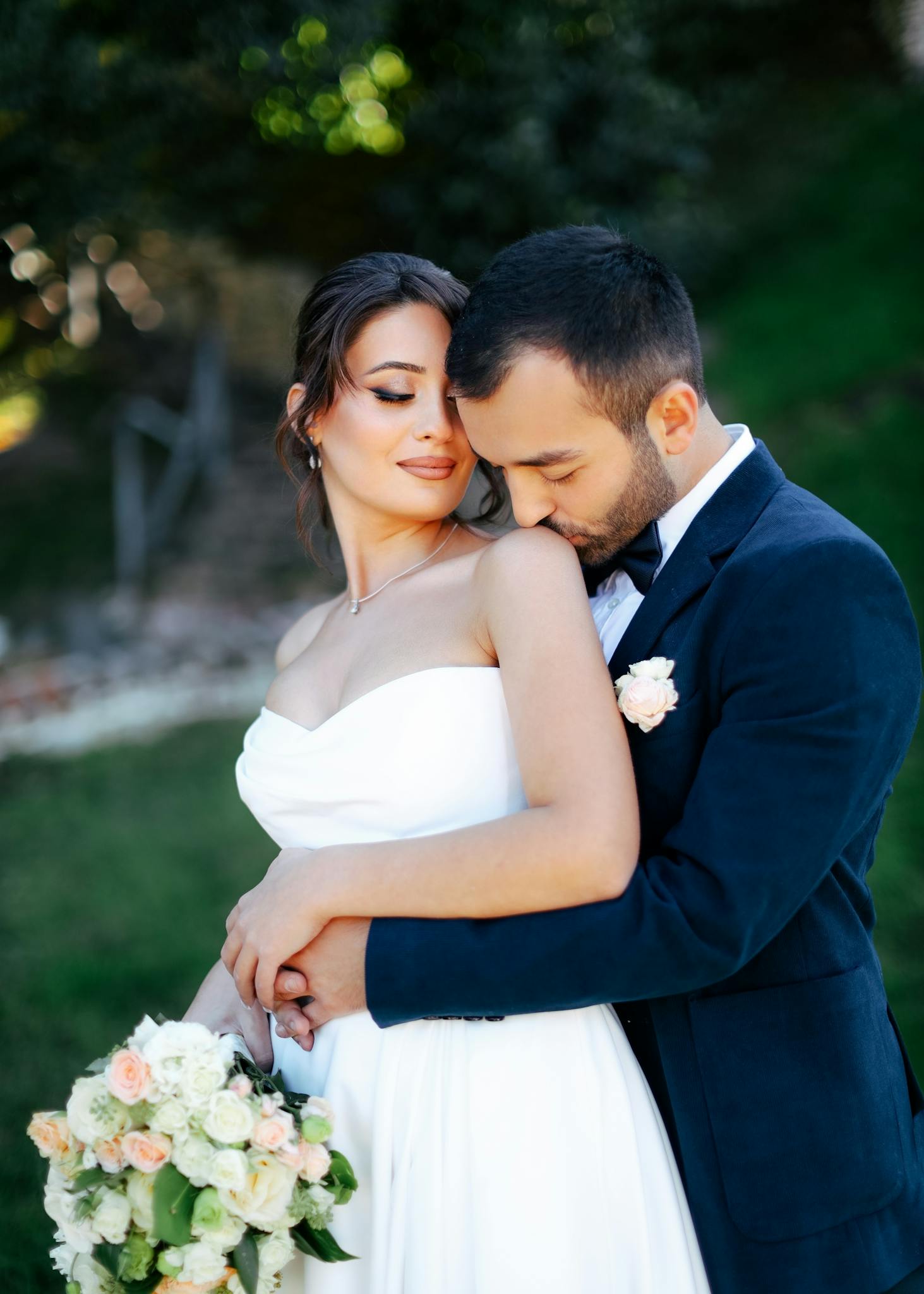 A bride and groom share a tender moment outdoors, captured in warm sunlight.