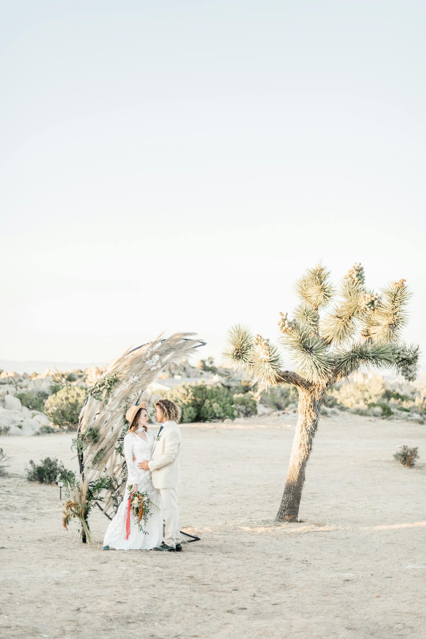 A couple in wedding attire embraces under a decorative arch in a serene desert landscape.